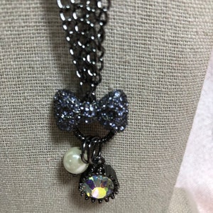 Può includere: Una collana grigio scuro con un ciondolo a forma di fiocco ricoperto di piccole pietre scintillanti. Sotto il fiocco, pendono una perla bianca e un ciondolo con una pietra ovale iridescente. La catena è fatta di maglie ad incastro.