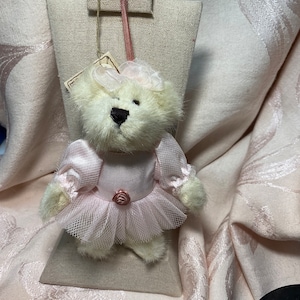 May include: A white plush ballerina bear wearing a pink tutu and a pink satin bodice with a pink rose accent. The bear is hanging from a beige fabric stand.