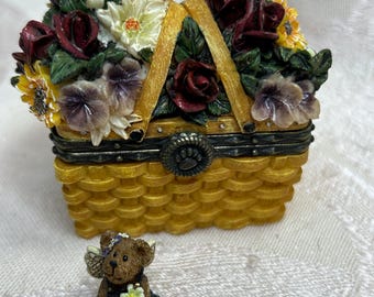 May include: A decorative yellow basket-shaped trinket box with a hinged lid, adorned with a floral arrangement of red roses, yellow sunflowers, and white flowers. A small teddy bear figurine sits nearby.