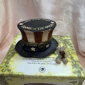 以下が含まれることがあります： 「Land of the Free, Home of the Brave」と書かれたミニチュアのアンクルサムの帽子が、装飾的な箱の上に置かれています。帽子は黒、白、赤で、金色の星が付いています。小さなテディベアの置物も箱の上にあります。
