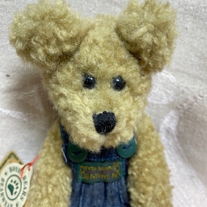 Op de afbeelding: Een beige pluche teddybeer met krullende vacht, zwarte plastic ogen en een zwart gestikte neus. De beer draagt een blauwe spijkerbroek met groene knopen en een label met de tekst Boyds Bears Bearwear. Een papieren label is aan de zijkant bevestigd.
