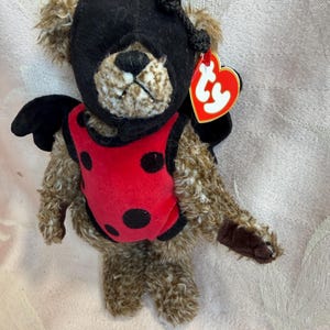 Può includere: Un orsacchiotto di peluche marrone che indossa un cappuccio nero e un gilet rosso a pois neri. L'orso ha ali nere attaccate alla schiena e un'etichetta Ty rossa a forma di cuore sull'orecchio. L'orso ha una pelliccia marrone e un naso nero.