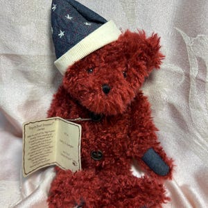 May include: A red teddy bear wearing a blue and white star patterned hat with a white brim. The bear has a blue denim patch on its right arm and a button on its chest. The bear is sitting on a pink surface. The tag reads "Boyds Best Dressed" series.