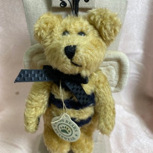 May include: A plush teddy bear with a bee-themed design. The bear has a yellow body with black stripes, a black bow, and white wings. A tag reads "Doris Bears Bearwear". The bear is displayed on a neutral-colored stand.
