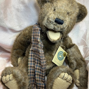 May include: A brown teddy bear with a plaid scarf. The bear has a black nose, tan paws, and a tag attached to its ear. The scarf is brown, blue, and tan. The bear is sitting on a pink surface.