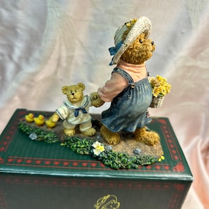 May include: A figurine of two bears, one wearing a sailor outfit and the other wearing a blue dress and a straw hat, walking together. The bears are holding hands and there are yellow chicks following behind them. The figurine is on a green base with flowers and is in a box with the words "Momma Mary's" on it.