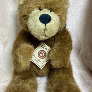 Puede incluir: Un oso de peluche marrón con ojos negros y nariz negra. El oso sostiene una etiqueta que dice "BOYD The Head Bean Collection".