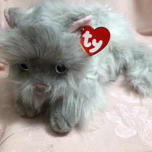 Pode incluir: Um brinquedo de pelúcia cinza com formato de gato com uma cauda longa e fofa e uma etiqueta vermelha em forma de coração. O gato tem o interior das orelhas rosa e uma textura macia e peluda. O brinquedo está sobre uma superfície rosa com estampa floral.