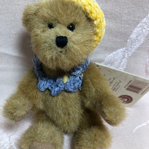 Könnte beinhalten: Ein kleiner brauner Teddybär, der einen gelben Häkelhut und einen blauen Häkelkragen trägt. Der Bär sitzt auf einer weißen Oberfläche.