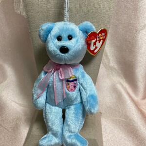 May include: A light blue Ty Beanie Baby with a pink ribbon around its neck. The bear has a small, embroidered patch on its chest that resembles an Easter egg. The bear is wearing a red Ty tag that says "Basket Beanies Ty".
