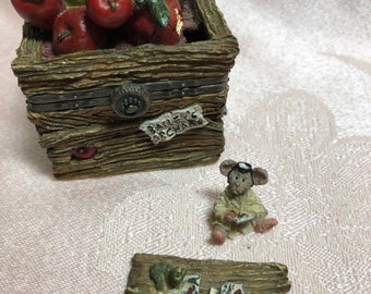 May include: A wooden box with a hinged lid, filled with red and green apples. The box has a label that reads "Bailey's Orchard". A small figurine of a mouse wearing a hat and holding a book sits on a wooden platform with apples and leaves.