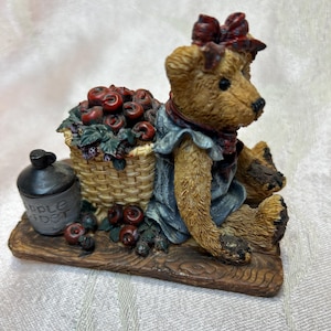 Puede incluir: Una figura de oso pardo con un vestido azul y un lazo rojo, sentada sobre una base de madera. El oso está al lado de una cesta de mimbre llena de manzanas rojas y una pequeña jarra negra con las palabras "Apple Cider".
