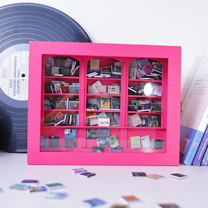 Może przedstawiać: Miniaturowa r&oacute;żowa biblioteczka z przeszklonymi drzwiami i p&oacute;łkami wypełnionymi małymi książkami. Biblioteczka stoi na białym tle, wok&oacute;ł niej rozrzucone są małe książeczki.