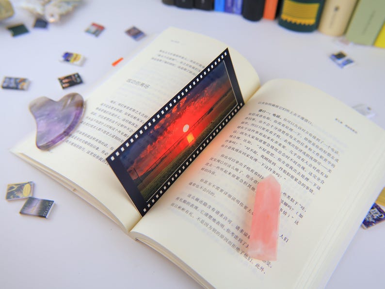May include: A book with a black and white film strip bookmark with a sunset photo. The bookmark is laying on a page with text in a foreign language. A purple heart-shaped stone is on the left page of the book.