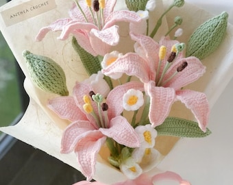 May include: A bouquet of crocheted pink lilies and white flowers wrapped in white paper with a pink ribbon. The bouquet is tied with a pink ribbon and has a tag that says "Anthea Crochet".