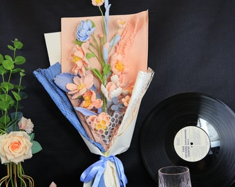 May include: A bouquet of crocheted flowers in shades of pink and blue, wrapped in white and pink tissue paper and tied with a blue ribbon. The bouquet is sitting on a black surface.
