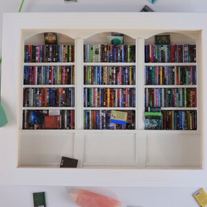 Może przedstawiać: Biała miniaturowa biblioteczka z trzema p&oacute;łkami wypełnionymi miniaturami książek. Biblioteczka znajduje się w białej ramie i stoi na białej powierzchni.