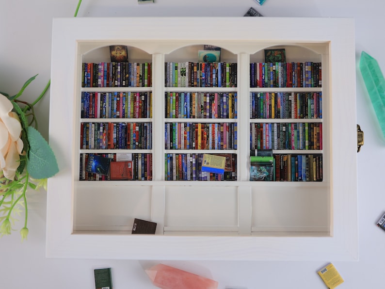 Może przedstawiać: Biała miniaturowa biblioteczka z trzema p&oacute;łkami wypełnionymi miniaturami książek. Biblioteczka znajduje się w białej ramie i stoi na białej powierzchni.
