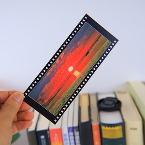 May include: A black and white film strip bookmark with a sunset scene. The sunset is orange, red, and yellow with a white sun in the center. The bookmark is on a stack of books.