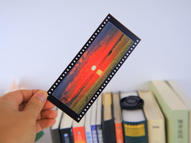 May include: A black and white film strip bookmark with a sunset scene. The sunset is orange, red, and yellow with a white sun in the center. The bookmark is on a stack of books.