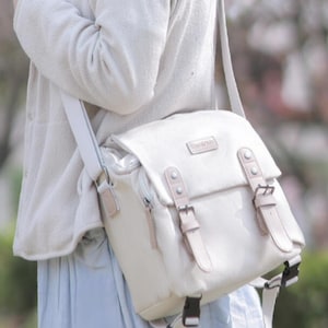 May include: Cream-colored shoulder bag with a flap closure and two buckle straps. The bag has a rectangular shape with a small, rectangular label on the front. The bag is being worn with a cream-colored sweater and a light blue skirt.