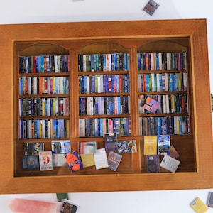 May include: A miniature wooden bookcase with glass doors and shelves filled with miniature books. The bookcase is brown and has a decorative top. There are several miniature books scattered around the bookcase.