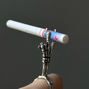 Könnte beinhalten: Ein silberner Skelett-Handring, der eine Zigarette hält. Die Zigarette hat einen weißen Filter und ein rosa-blaues Design. Der Ring hat knöcherne Finger und ein rundes Band.