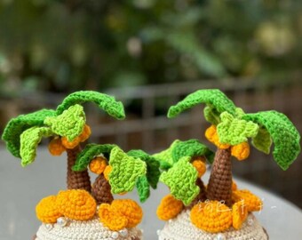 May include: Two crocheted palm trees with green leaves, brown trunks, and orange fruit. The trees sit on a brown base with a white and tan trim, adorned with small white beads. The background is a blurred green.