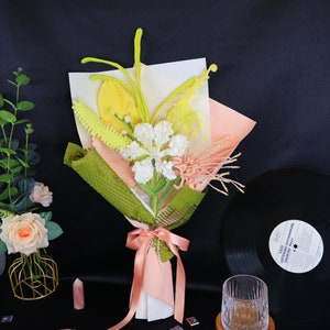 May include: A bouquet of artificial flowers wrapped in green and pink tissue paper. The bouquet includes yellow, white, and pink flowers. The bouquet is tied with a pink ribbon.