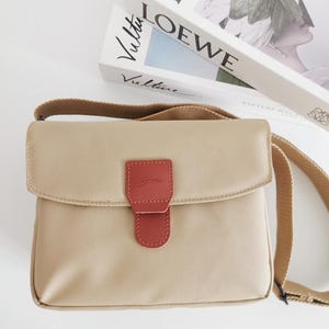 Peut inclure: Un sac bandoulière beige avec une fermeture à rabat et un accent en cuir rouge. Le sac a une sangle beige et une petite languette en cuir rouge avec du texte. Le sac est posé sur une surface blanche.