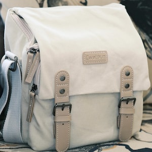 May include: Cream canvas messenger bag with a flap closure and adjustable shoulder strap. Features beige leather accents, including straps with buckles and a small rectangular patch with the text "Cwatsun". The bag has a zippered main compartment.