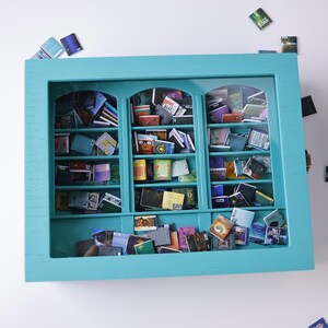Może przedstawiać: Miniaturowa biblioteczka w kolorze turkusowym z przeszklonymi drzwiami i p&oacute;łkami wypełnionymi miniaturami książek. Biblioteczka ma szufladę na dole.