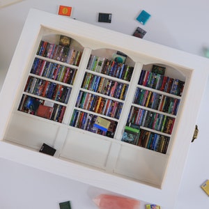 Może przedstawiać: Biała miniaturowa biblioteczka z trzema sekcjami, każda wypełniona kolorowymi miniaturami książek. Biblioteczka jest oprawiona w białą ramkę i ma ozdobny łuk na g&oacute;rze każdej sekcji.