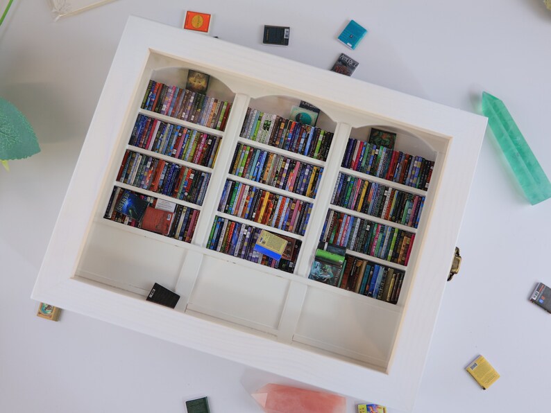 Może przedstawiać: Biała miniaturowa biblioteczka z trzema sekcjami, każda wypełniona kolorowymi miniaturami książek. Biblioteczka jest oprawiona w białą ramkę i ma ozdobny łuk na g&oacute;rze każdej sekcji.