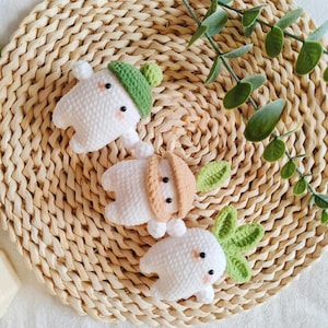 May include: Three handmade crochet radish toys with white bodies, green and tan accents, and black eyes. The toys are arranged on a woven straw mat with green leafy sprigs.
