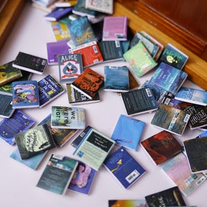 Peut inclure: Une collection de livres miniatures aux couvertures colorées, dispersés sur une surface blanche. Les livres présentent divers titres et motifs, dont "Alice" et "Coraline". La scène évoque un sentiment de fantaisie et de charme littéraire.