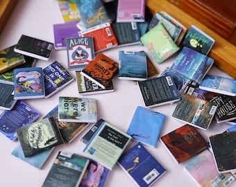 May include: A collection of miniature books with colorful covers scattered on a white surface. The books have various titles and designs, including "Alice," "Coraline," and "The Ghost." The scene evokes a sense of whimsy and literary charm.
