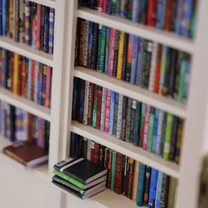 Może przedstawiać: Biała biblioteczka wypełniona kolorowymi książkami. Książki są uporządkowane według koloru i rozmiaru. Biblioteczka jest wykonana z drewna i ma prosty design.