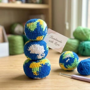 May include: Three blue, yellow, and green crocheted globe ornaments stacked on a wooden surface. A fourth globe and a ball of blue yarn are next to the stack. A small card reads "Handmade with Love".