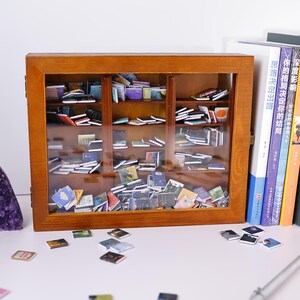 Może przedstawiać: Miniaturowa drewniana biblioteczka wypełniona małymi książkami. Biblioteczka ma szklane drzwi i stoi na białej powierzchni. Kilka małych książek jest rozrzuconych na powierzchni przed biblioteczką.