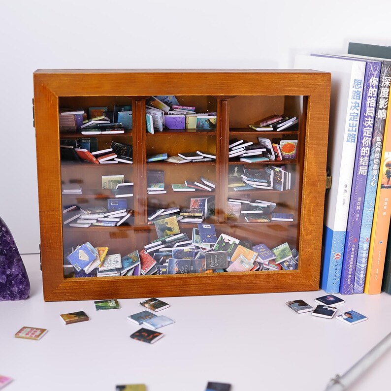 Może przedstawiać: Miniaturowa drewniana biblioteczka wypełniona małymi książkami. Biblioteczka ma szklane drzwi i stoi na białej powierzchni. Kilka małych książek jest rozrzuconych na powierzchni przed biblioteczką.