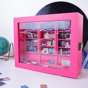 Może przedstawiać: R&oacute;żowa drewniana biblioteczka z przeszklonymi drzwiami i p&oacute;łkami wypełnionymi miniaturowymi książkami. Biblioteczka stoi na białym blacie.