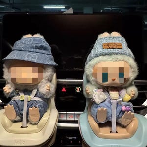 Peut inclure: Deux poupées dans des sièges auto. Les poupées portent des tenues et des chapeaux en jean. Les sièges auto sont beiges et bleus. Les poupées ont des bordures en fourrure autour de leur visage. Les sièges auto ont le mot "hello".