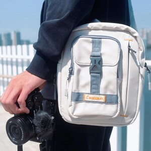 May include: A light gray messenger bag with a blue strap and accents. The bag has a front pocket with a buckle closure and the text "Cwataun" on the front. A black camera with a microphone is held in the hand.