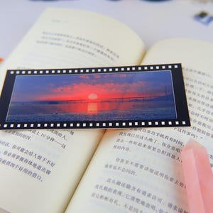 May include: A black and white film strip bookmark with a colorful sunset over a bridge and water. The bookmark is lying open on a book with a pink crystal on top of the book.
