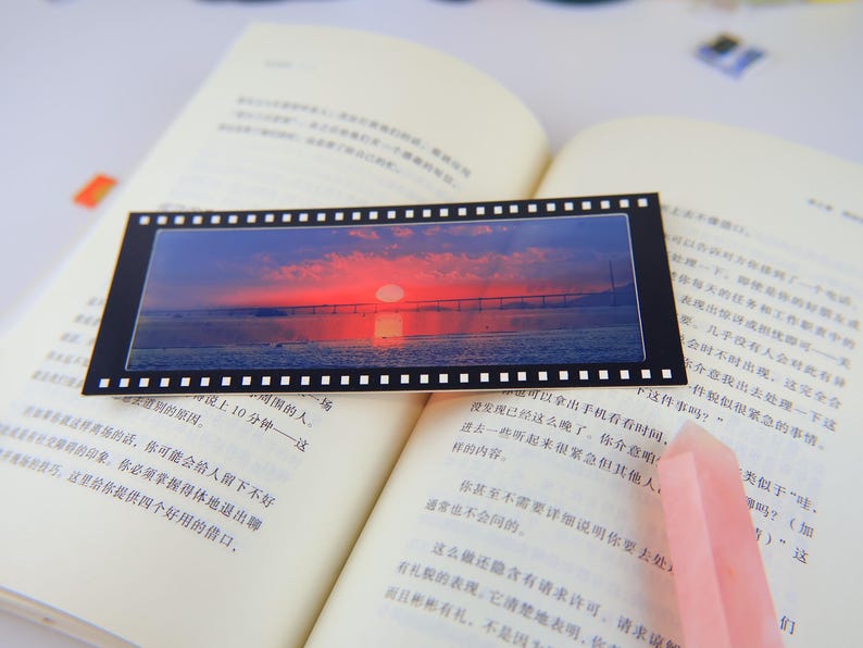 May include: A black and white film strip bookmark with a colorful sunset over a bridge and water. The bookmark is lying open on a book with a pink crystal on top of the book.