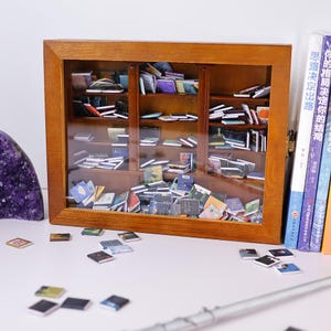 Może przedstawiać: Miniaturowa drewniana biblioteczka wypełniona małymi książkami. Biblioteczka ma szklane drzwi i stoi na białym tle. Wok&oacute;ł biblioteczki rozrzucone są małe książki.