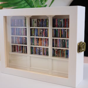 May include: A white wooden frame with a glass front showcasing a miniature bookshelf filled with colorful books. The bookshelf has three sections with multiple shelves, each filled with miniature books.