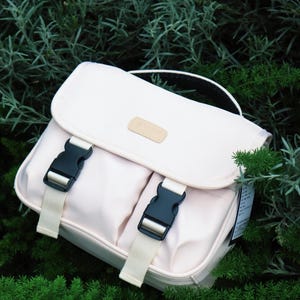 May include: A light pink satchel bag with a top handle and two front pockets. The bag features black buckles with cream-colored straps and a small tan patch on the front. The bag is set against a backdrop of green foliage.