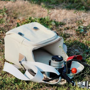 May include: A cream-coloured canvas camera bag with a matching shoulder strap, open to reveal a black camera with a lens cap. A red camera strap and a small red tomato are also visible inside the bag, resting on green grass.
